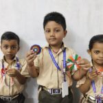 Three schoolchildren in beige uniforms hold colorful handmade badges posing against a white wall   center child shows a blue and purple rosette left and right children display other crafts all with lanyards